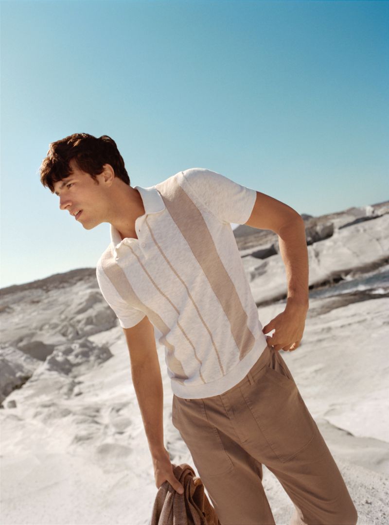Man wearing a beige polo shirt and light brown pants standing on a rock formation with a clear sky in the background, fashion shoot.