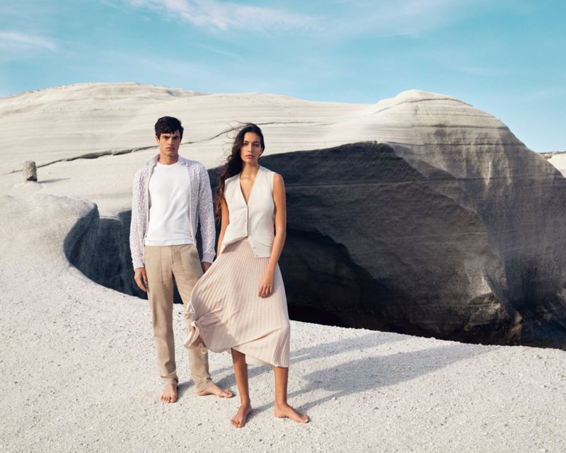 Two models, a man and a woman, pose barefoot in light-colored clothing against a backdrop of unusual rock formations and a blue sky.