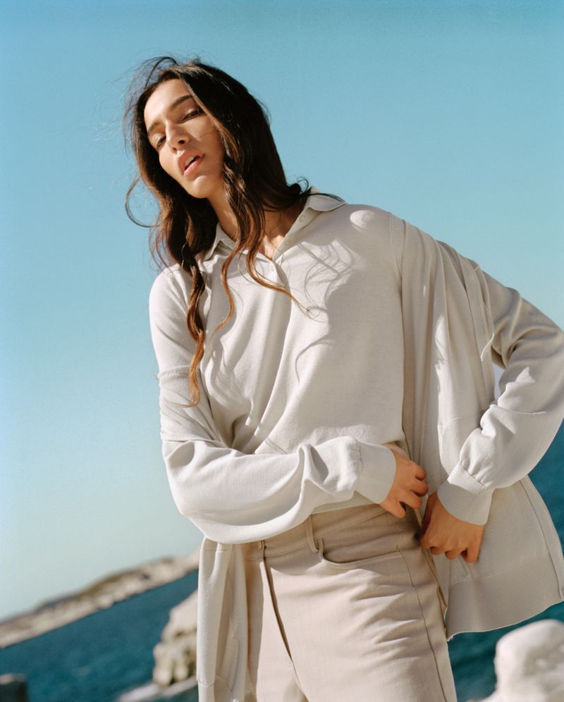 A woman poses in a cream-colored outfit with long hair blowing in the wind against a light blue sky and ocean backdrop.