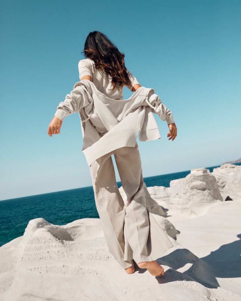 Woman in linen suit standing on white rock formation by ocean under blue sky, wind blowing jacket open, back to camera.