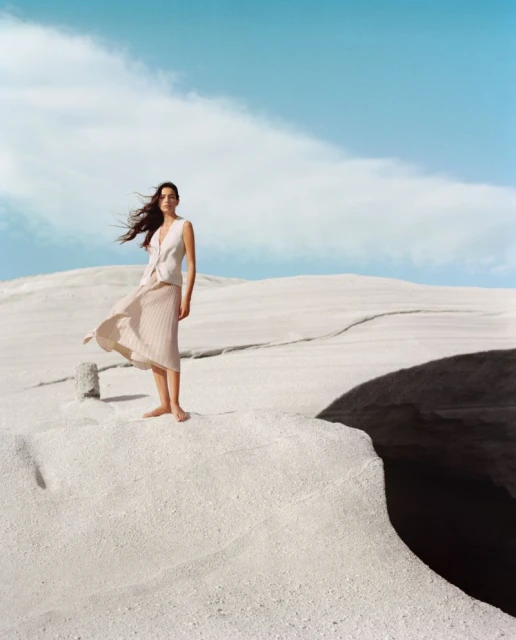 An adult female stands barefoot in a flowing skirt and sleeveless top on a white landscape with a clear sky in the background.