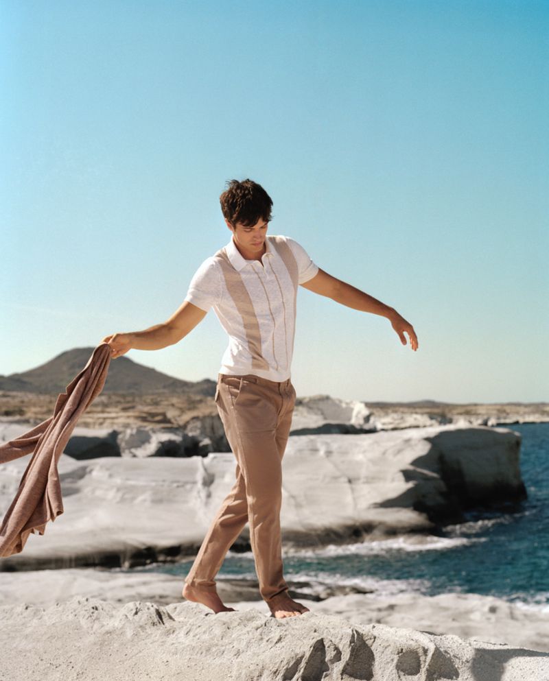 Young adult male walks barefoot on a rocky shoreline holding a brown jacket, set against a blue sky and white rock formations.