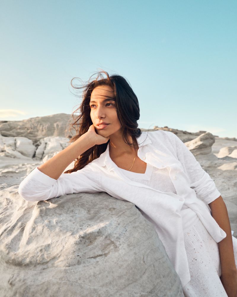 Woman in white leans on rock formation outdoors with blue sky. Female with long brown hair poses in flowing white clothing and jewelry.