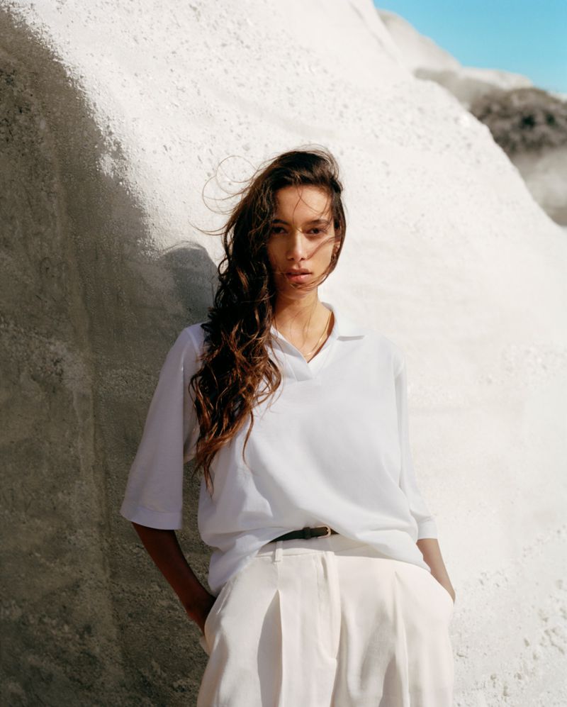 Brunette woman in white shirt poses outdoors against white rock formation on a sunny day. Fashion and travel concept.