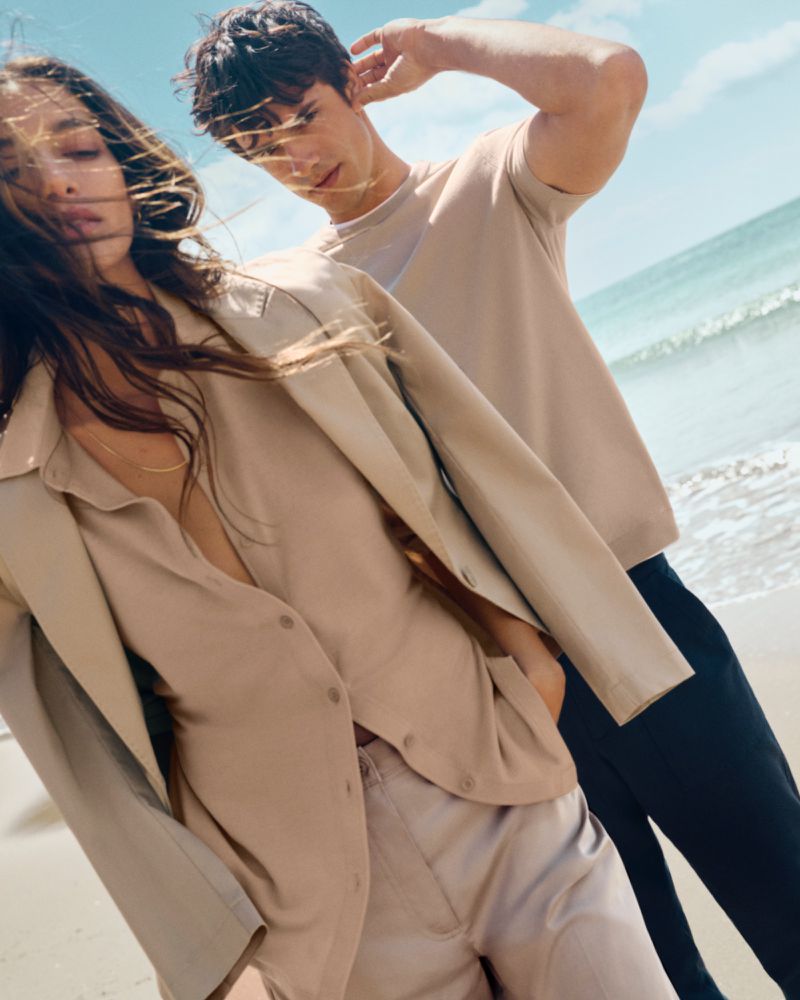 Two people pose on a beach wearing beige clothing. The woman is in the foreground, with ocean and sky in the background on a sunny day.