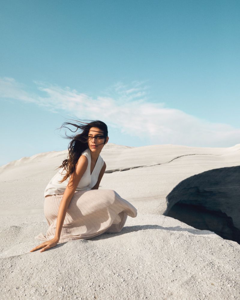 A woman in a white sleeveless top and beige skirt is sitting on a sandy surface with a blue sky and clouds behind her.
