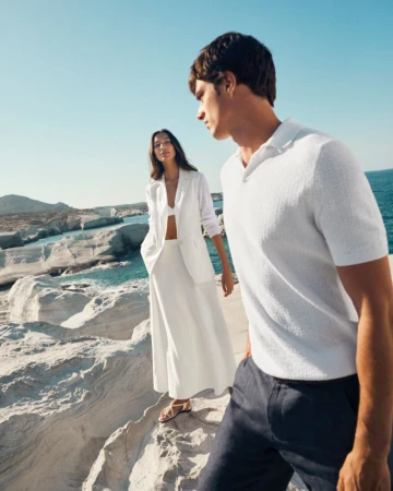 Fashion shoot of a man and woman in white summer outfits standing on white rocks overlooking the sea on a sunny day.