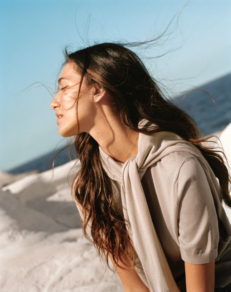 Profile view of a young woman with long dark hair blowing in the wind, wearing a beige top, with a light blue sky in the background.