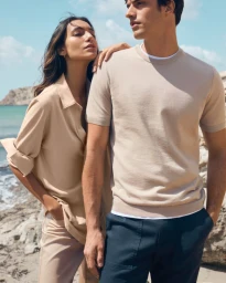 A man and woman in neutral knitwear pose outdoors near the coast with a blue sea and rocky background under a bright sky.