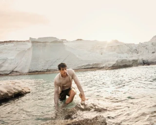 Man wading in coastal shallow water wearing a beige shirt against pale rock formations under a soft sky