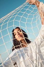 Model wearing a white shirt looking through a knotted rope net against a clear blue sky background
