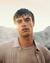Portrait of a man with wet hair wearing a beige button-up shirt against a mountain backdrop under natural backlighting.