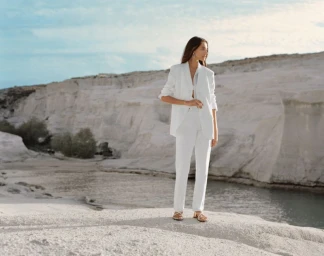 Model standing on a pale rock formation wearing a white blazer and white trousers near water.