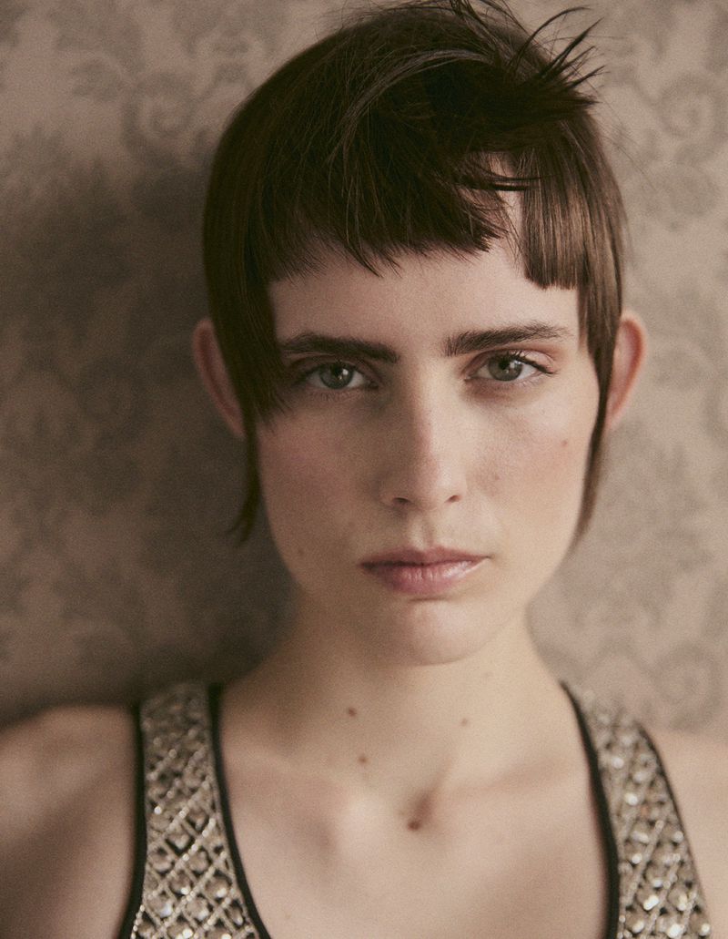 Close-up portrait of a young adult woman with short brown hair and an embellished top, set against a muted patterned background.