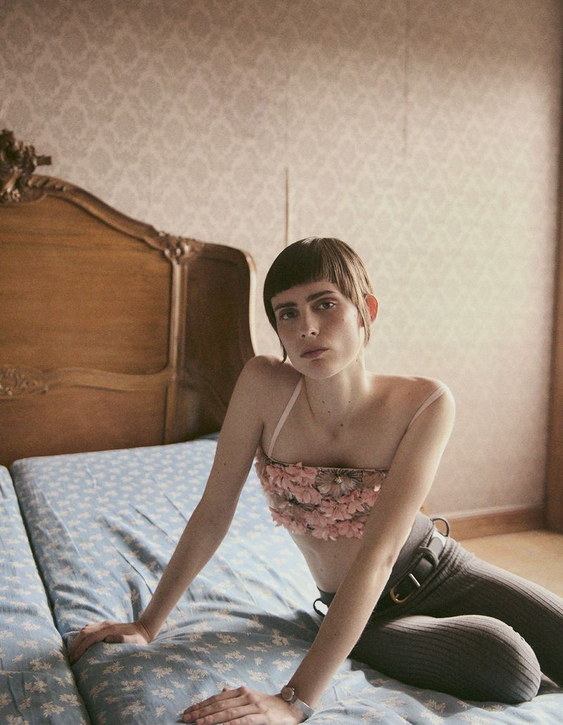 A slender brunette woman poses on a floral-patterned bed wearing a pink flower top, gray pants, and a wristwatch in soft, diffused light.