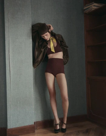 A fashion model poses indoors in a maroon two-piece outfit, a yellow neck scarf, and a brown leather jacket against a blue wall.