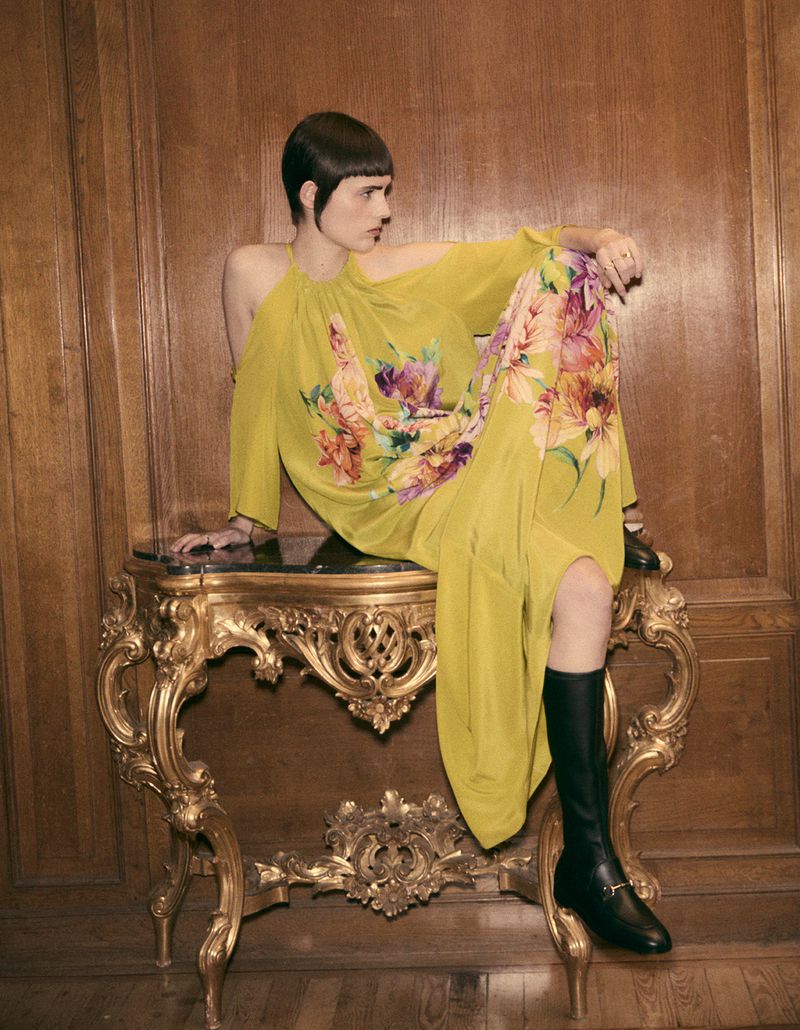 A stylish woman sits on an ornate gold table in a yellow floral dress and black boots against a paneled wood background.