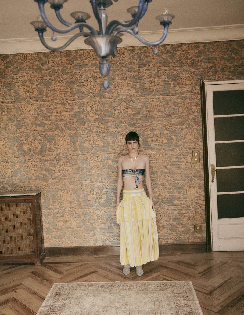 A young adult woman stands in a room with patterned wallpaper, parquet floors, and a rug, wearing a two-piece outfit and accessorized with a necklace.