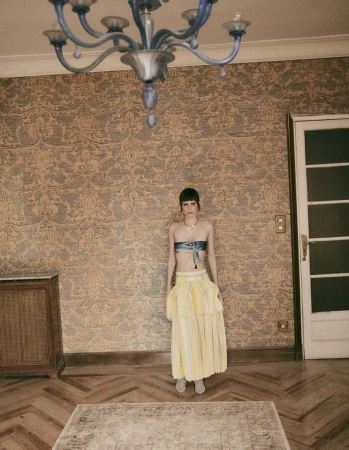 A young adult woman stands in a room with patterned wallpaper, parquet floors, and a rug, wearing a two-piece outfit and accessorized with a necklace.