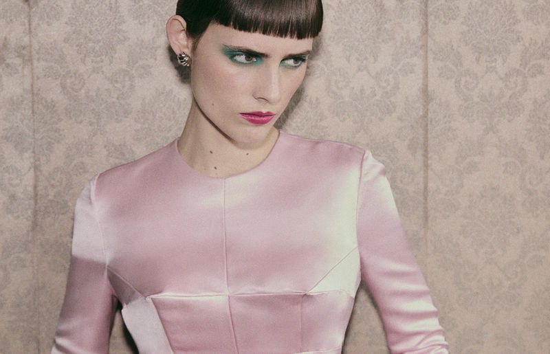 Close-up studio portrait of a woman with a short, dark hairstyle, green eyeshadow, pink lipstick, and a light pink satin dress.