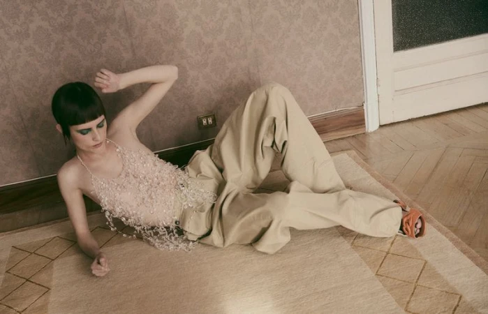 A fashion shot of a young adult female model with dark, short hair wearing a beaded top and loose pants, reclining on a carpeted floor.