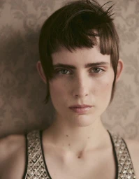 Close-up portrait of model Estrella Gomez with short textured hair and patterned top against a damask patterned wall.