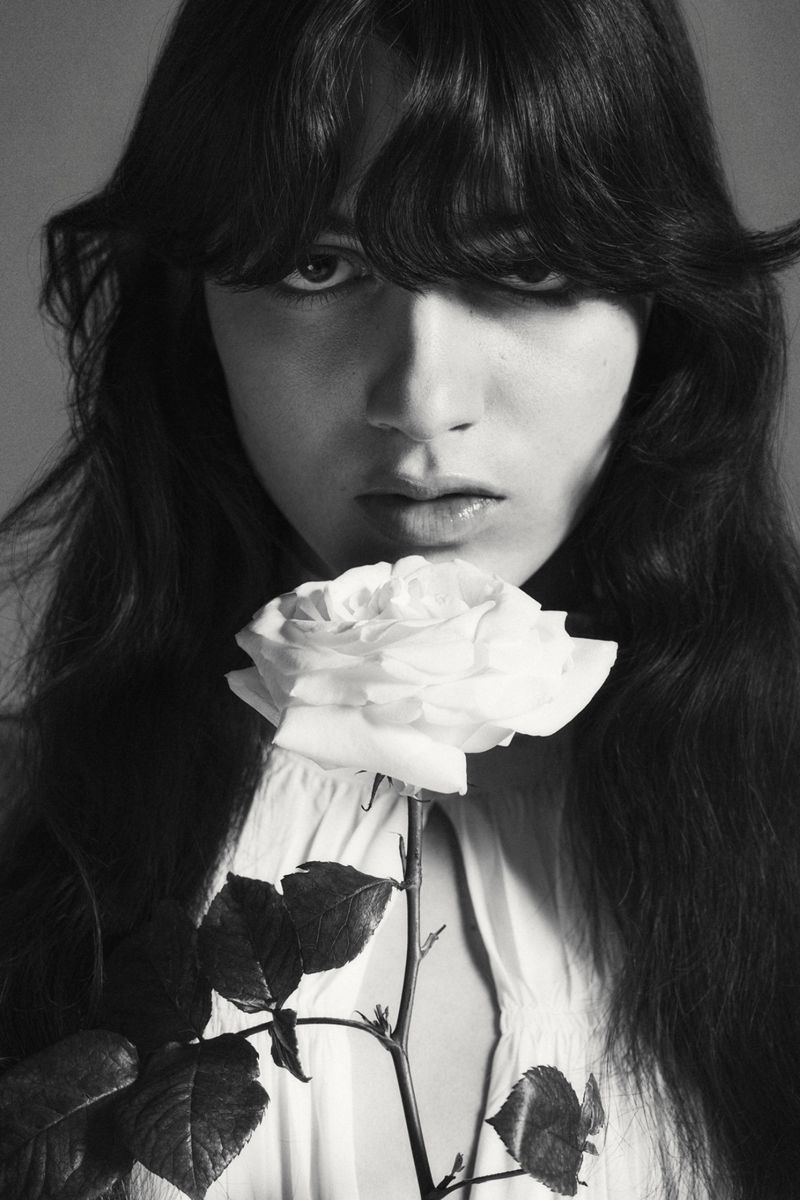 Monochrome portrait of a young woman with long hair holding a white rose. Close-up shot with a blurred background in a studio setting.