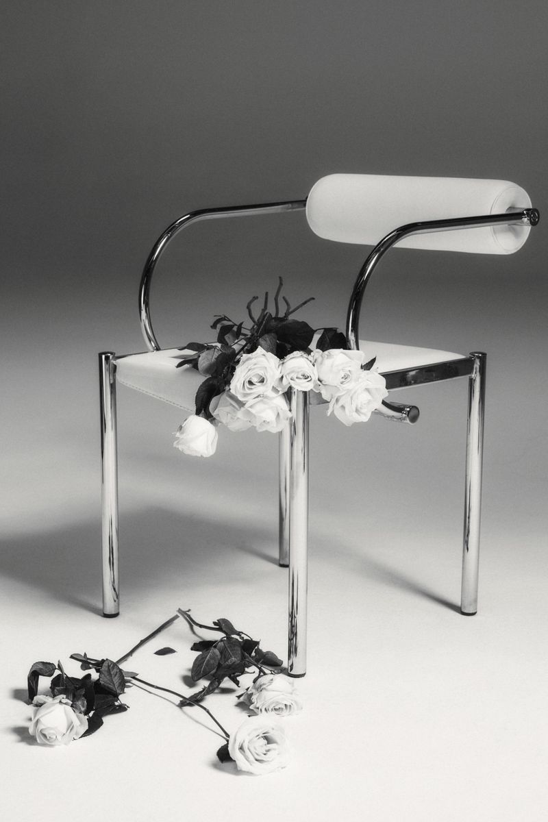 Arrangement of white roses on and around a chrome chair against a neutral backdrop, creating a minimalist still life.