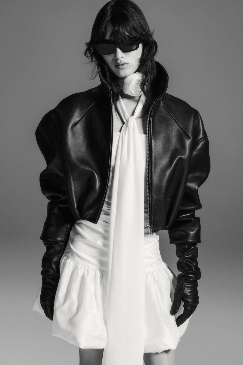 Model in black leather jacket and sunglasses wearing a white dress against grey seamless background
