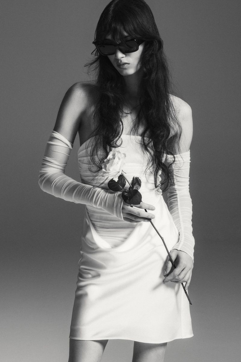 Model in white strapless dress wearing sunglasses holding a single rose against a grey studio backdrop.