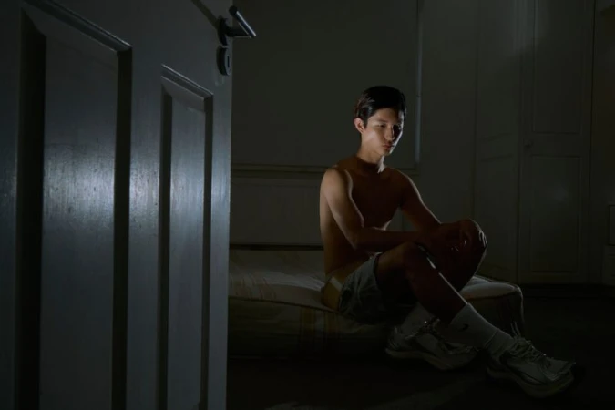 Young adult sits on a bed in a dimly lit room with an open door, highlighting the mood and simple setting.