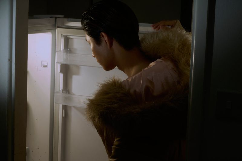 Adult male looks into an empty refrigerator in an indoor setting with neutral tones, soft lighting, and a fur-lined coat.