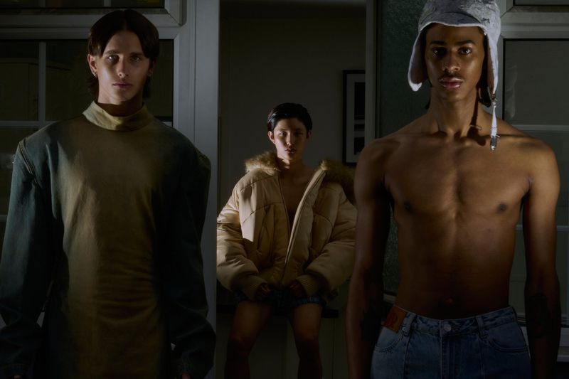 Three models pose indoors in a darkly lit room. Two are shirtless, wearing jeans and a cap, while another is wearing a jacket.