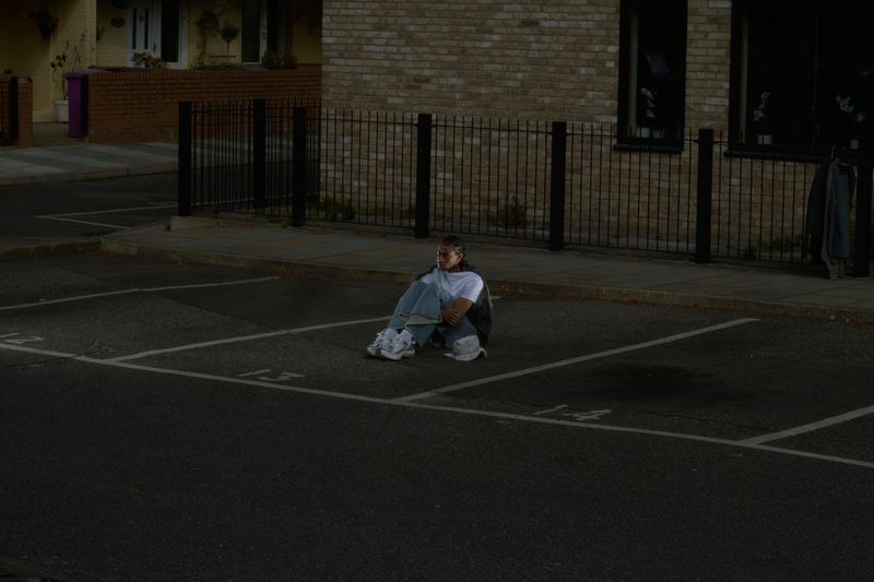 Person sits on empty parking lot at night with street lights. Wearing jeans and white sneakers in the dark urban setting.