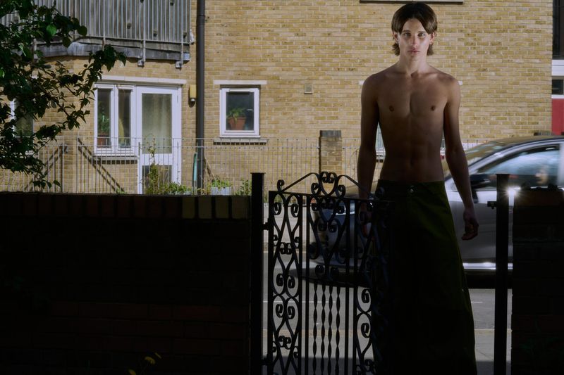 Adult male stands near an ornate gate with a brick building and car in the background, wearing a green wrap.
