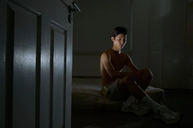 Young man sitting on a low mattress in a dark room illuminated by a single soft light source