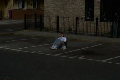 A person sitting on the asphalt in a marked parking space in front of a brick building exterior.