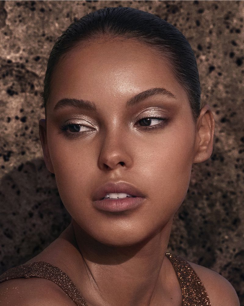 Close-up portrait of a woman with brown skin and neutral makeup, looking off-camera, set against a textured brown backdrop.