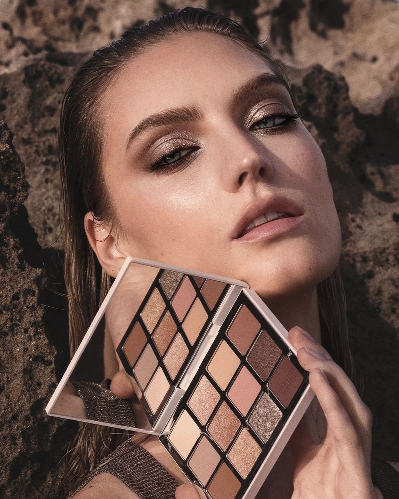 A close-up shot of a woman with smokey eyeshadow and neutral lipstick, holding an eyeshadow palette with both hands. Rock formations behind.