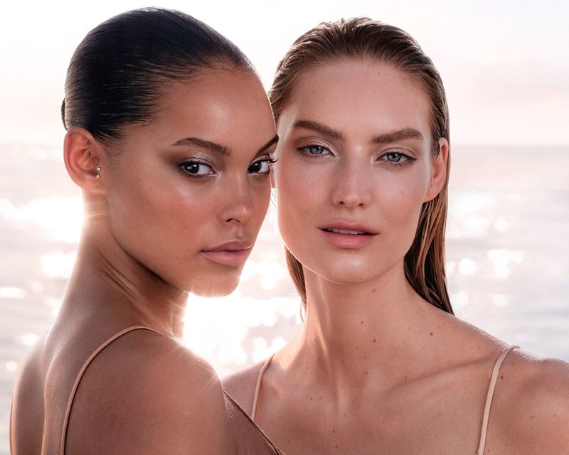 Two women pose closely together with natural makeup in an outdoor setting with blurred sunlight background. Beauty and fashion imagery.