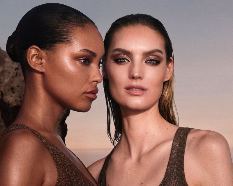 Two women pose in metallic bronze tops against a softly lit outdoor backdrop; beauty and fashion portrait featuring natural, dewy makeup.