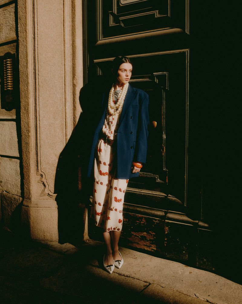 Fashionable woman in double-breasted blazer, printed dress, and pearl necklace poses against a dark door on a sunny urban street.