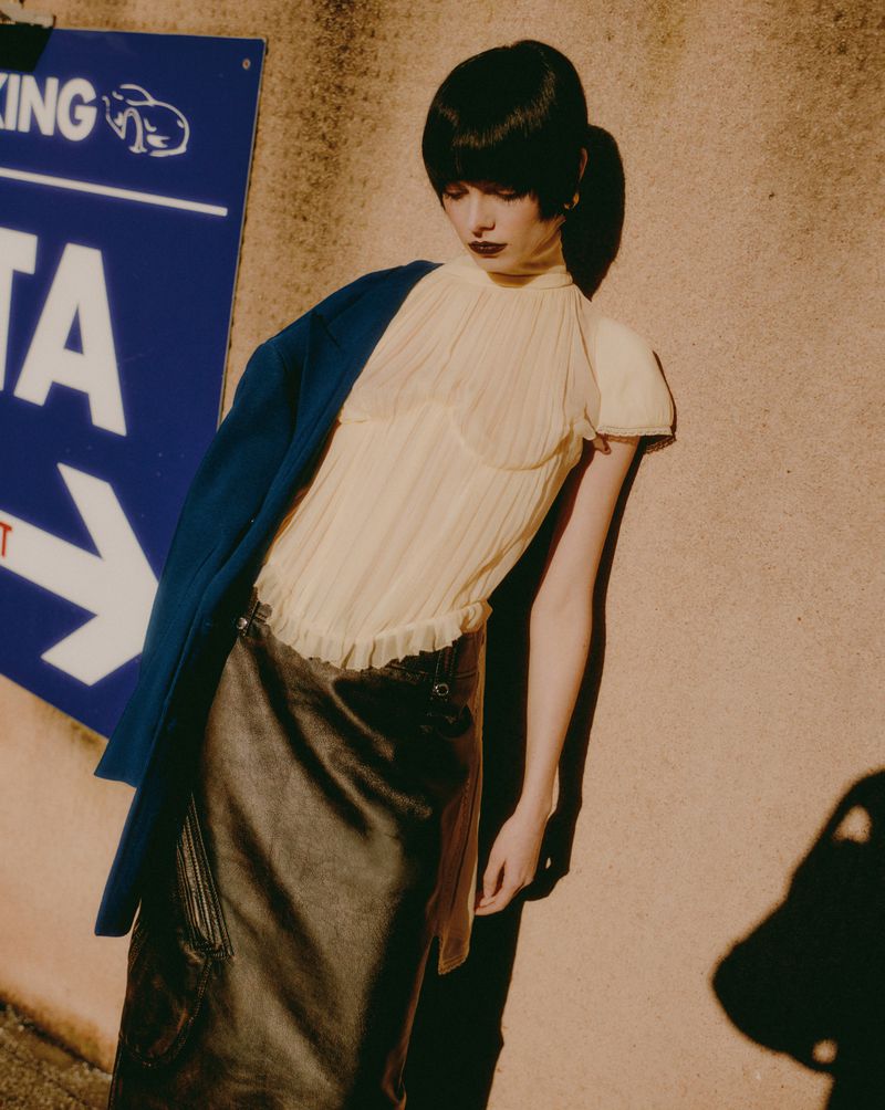 A fair-skinned woman with a black bob stands against a neutral wall, wearing a yellow blouse, black skirt, blue jacket, and dark lipstick.