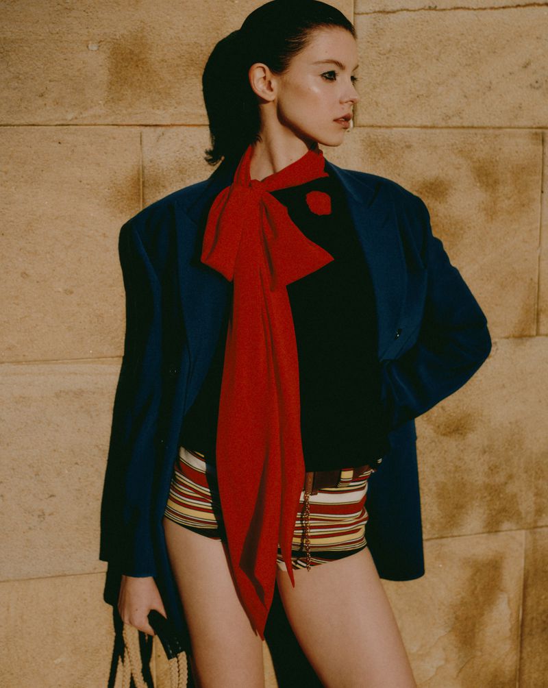 Portrait of a woman in a blue blazer and striped shorts standing against a textured wall. She wears a long red scarf and holds a bag.