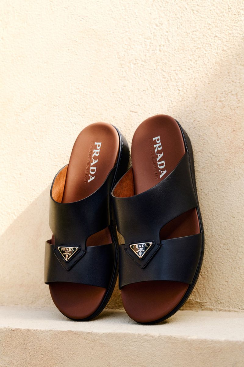 Prada slide sandals in black leather with brown insoles are displayed indoors with a white wall backdrop, showcasing luxury footwear.