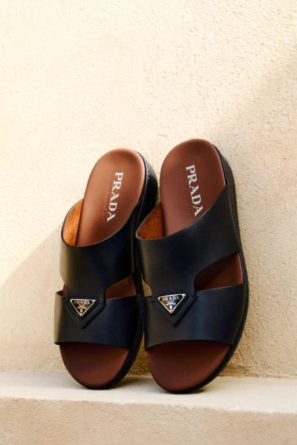 Prada slide sandals in black leather with brown insoles are displayed indoors with a white wall backdrop, showcasing luxury footwear.