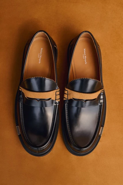 Maison Margiela black leather penny loafers with a tan suede strap, presented on a solid tan background in a top-down studio product shot.