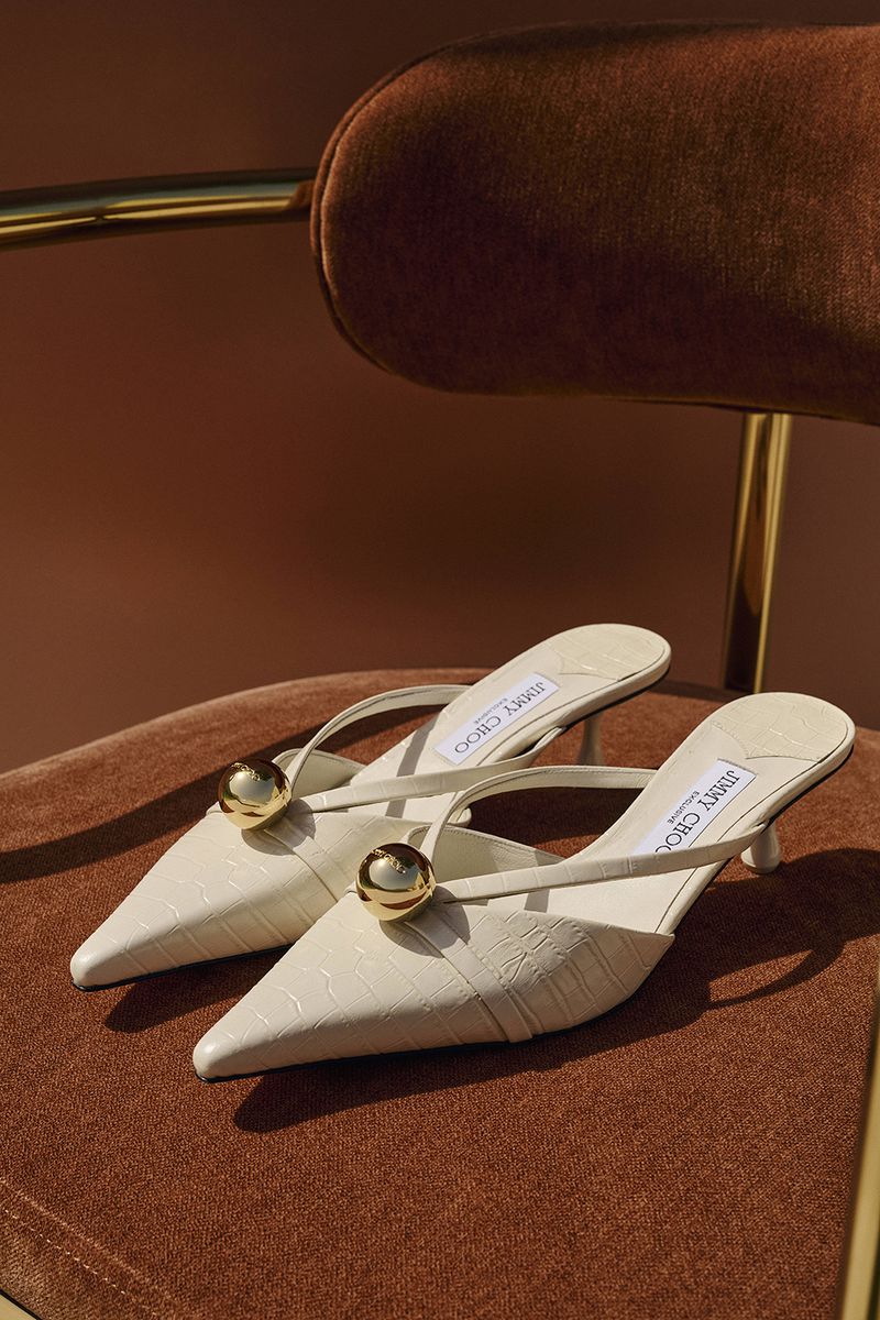 Cream Jimmy Choo mule heels with gold sphere embellishments on a brown velvet chair, fashion footwear, indoor product shot.