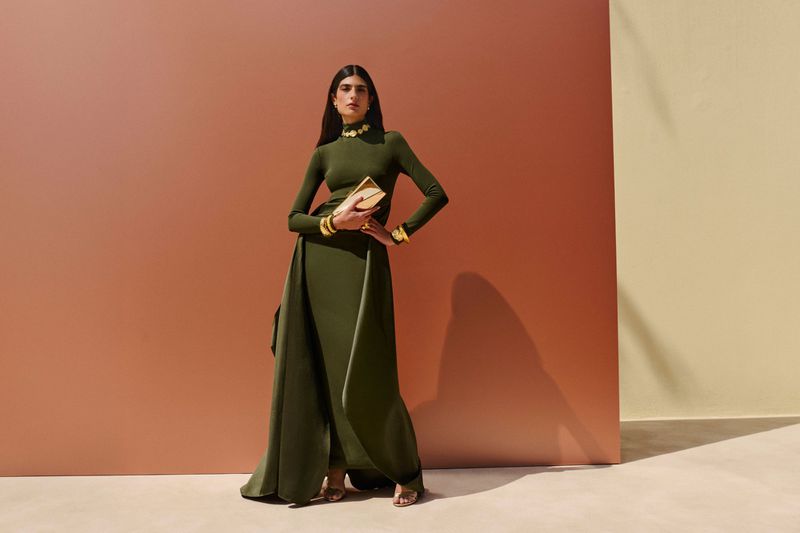 Elegant woman in long-sleeve green dress with gold jewelry stands holding gold clutch in a fashion studio against a warm background.