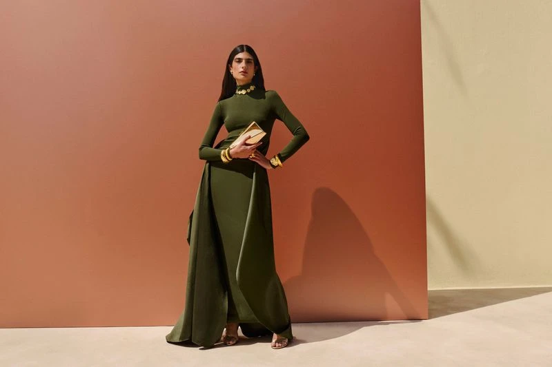 Elegant woman in long-sleeve green dress with gold jewelry stands holding gold clutch in a fashion studio against a warm background.